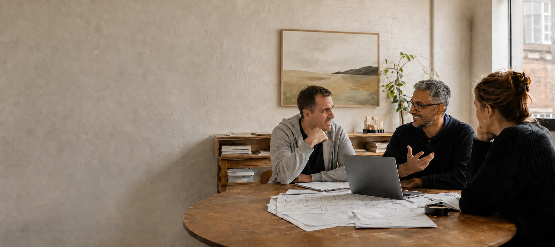 Three colleagues talking around a table covered in floor plans, with a laptop open between them